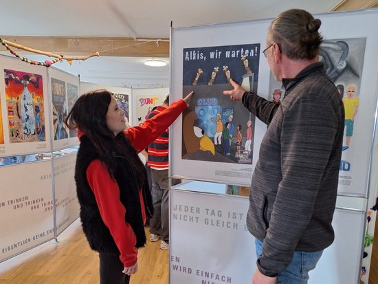 Ein Mann und eine Frau stehen in einer Ausstellung vor einem Posterstand. Beide zeigen auf ein Plakat mit der Überschrift „Albis, wir warten!“. Darunter steht „Jeder Tag ist nicht gleich“. Im Hintergrund sind weitere Tafeln und Lichterketten sichtbar.