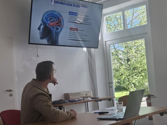 Ein Mann sitzt an einem Tisch vor einem Laptop und betrachtet einen großen Bildschirm. Dieser zeigt eine Präsentation mit dem Titel „Neurologie" und einer Illustration eines menschlichen Kopfes mit einem leuchtenden Gehirn.