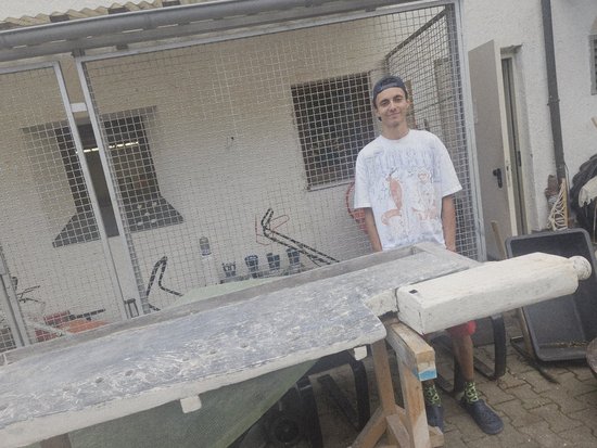 Ein junger Mann steht im Freien hinter einer großen, flachen Betonplatte und hält ein weißes zylindrisches Objekt darauf. Im Hintergrund ist ein Drahtgehege zu sehen.