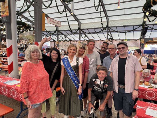 Eine Gruppe von Menschen posiert in einem Festzelt. In der Mitte steht eine Frau in Tracht mit einem blauen Band, auf dem „Schönste Wiesnwirtin“ steht. Sie wird von Freunden und Bekannten umringt, die alle in die Kamera lächeln.