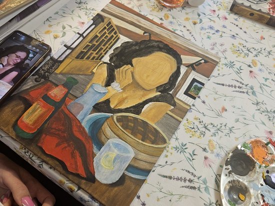 Ein Gemälde einer Frau liegt auf einer Tischdecke mit Blumenmuster. Daneben befinden sich eine Farbpalette, ein Smartphone mit Referenzfoto und zwei Gläser mit Getränk.