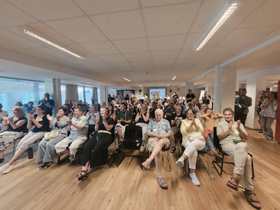 Eine große Gruppe von Menschen sitzt in Reihen in einem hellen Raum und applaudiert. Sie blicken in dieselbe Richtung und klatschen Beifall.