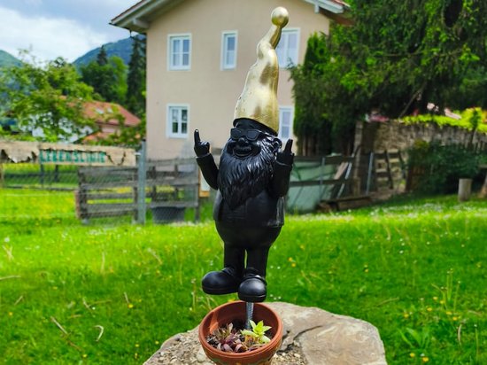 Ein schwarzer Gartenzwerg balanciert einen goldenen Ball auf der Nase und steht auf einem Turm aus fünf mit Sukkulenten bepflanzten Blumentöpfen, die auf einem großen Stein im Gras stehen.