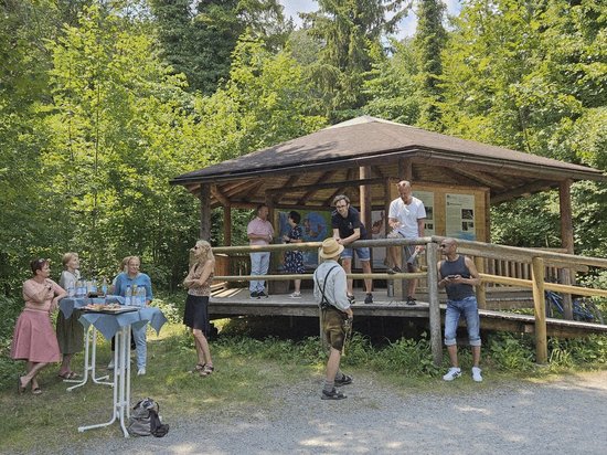 Eine Gruppe von Menschen versammelt sich im Wald um einen hölzernen Pavillon. Einige stehen auf der Veranda, andere auf dem Kiesweg daneben. Links steht ein Tisch mit Getränken. Die Szene wirkt wie eine entspannte Pause bei schönem Wetter.