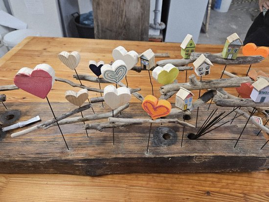 Ein rustikales Holzbrett mit Treibholz, in das zahlreiche kleine, bemalte Holzhäuschen und Herzen auf Stäbchen gesteckt sind.