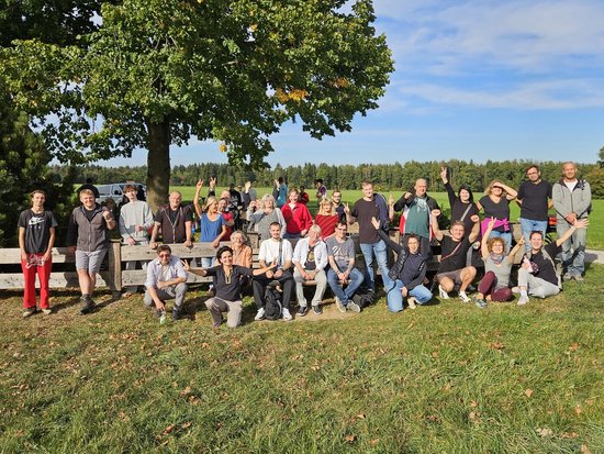 Eine große Gruppe von Menschen posiert fröhlich für ein Gruppenfoto auf einer Wiese unter einem großen Baum an einem sonnigen Tag.