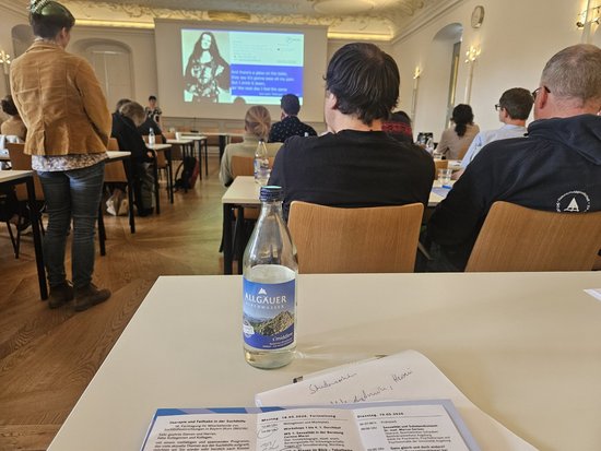 Teilnehmer sitzen in einem hellen Saal und hören einer Präsentation auf einer Leinwand zu. Im Vordergrund befindet sich ein Tisch mit einer Wasserflasche und ausliegenden Dokumenten.