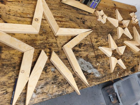 Auf einer Holzwerkbank sind verschiedene hölzerne Sternformen zu sehen. Neben großen, flachen Sternrahmen liegen mehrere kleinere, dreidimensionale geometrische Sterne, die aus einzelnen Holzstücken zusammengesetzt sind.