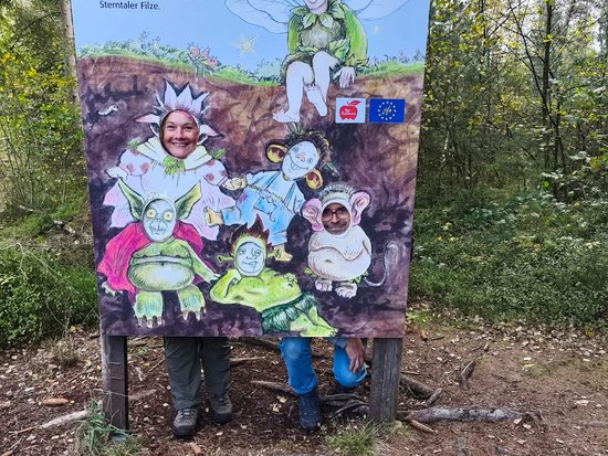 Ein großes, bunt bemaltes Holzschild im Wald zeigt fantasievolle Wesen wie Gnomen und Elfen. Zwei Personen stehen dahinter und posieren, wobei nur ihre Beine zu sehen sind. Oben sind Logos und ein Schriftzug erkennbar.